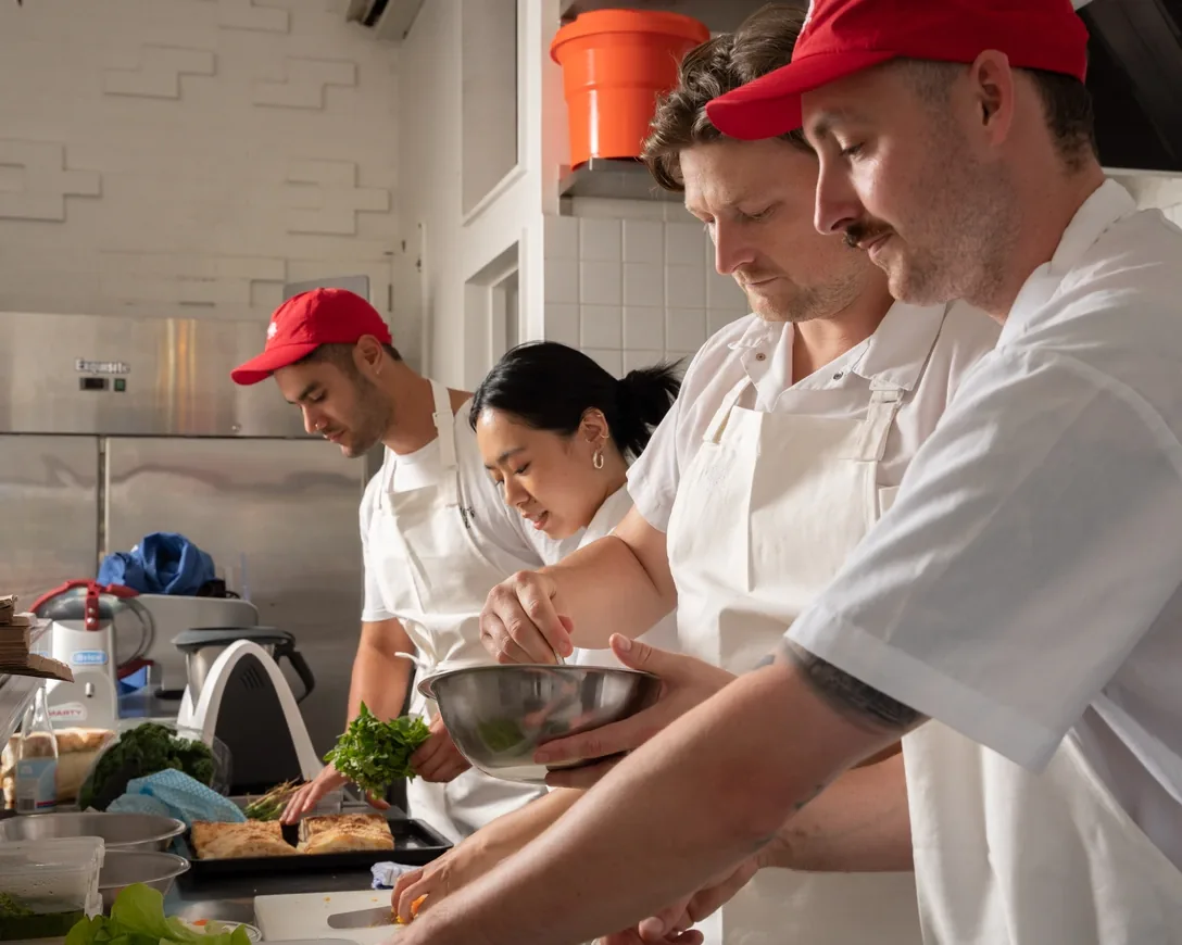 Chefs in red caps prepping greens and pastries side by side in a kitchen