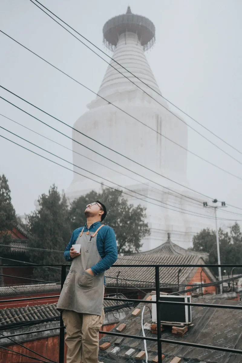 A barista with a cup, looking up from a rooftop beneath a white pagoda in Beijing