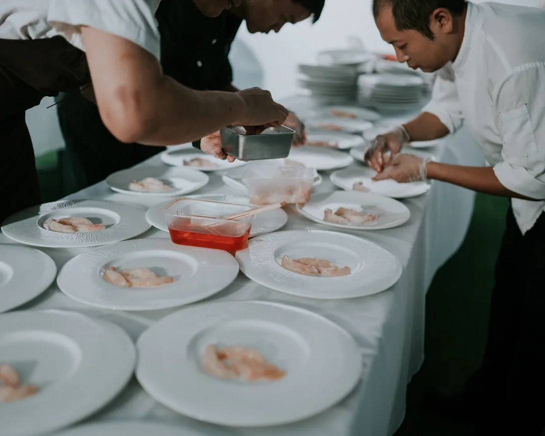 Chefs plating raw fish onto rows of white dishes at a long table