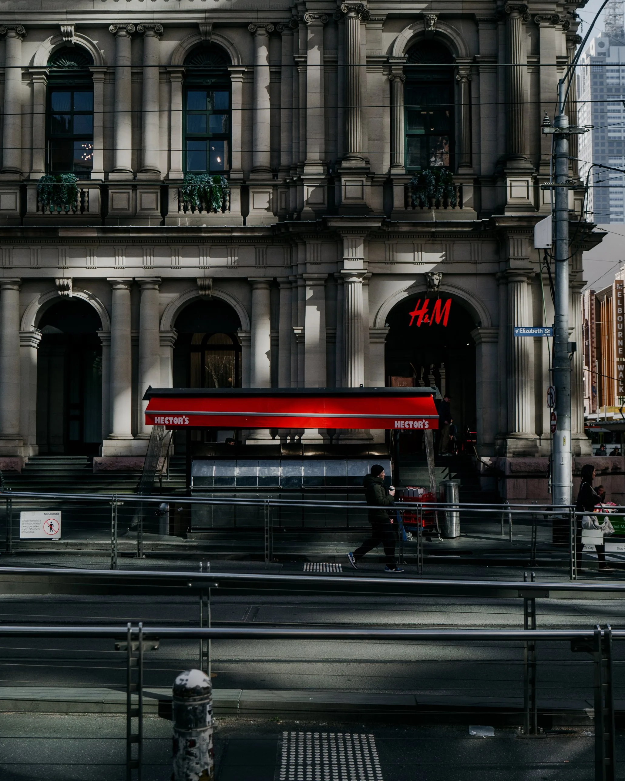 Hector’s Deli’s red-awninged kiosk on Elizabeth Street, Melbourne, in shafts of light beneath an ornate stone facade