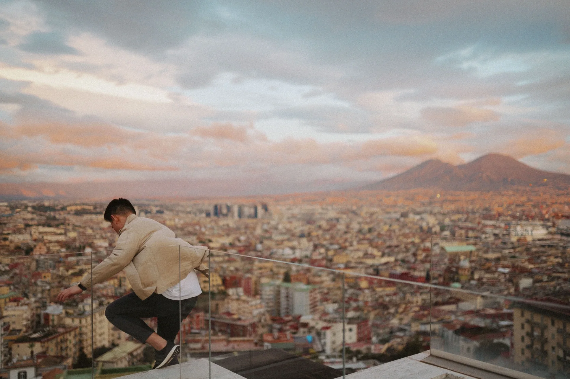 Weiming climbing onto a rooftop ledge above Naples at dusk, Mount Vesuvius on the horizon